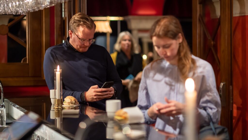 Två personer, en man och en ung kvinna, står vid en bardisk, båda tittar ner på sina mobiler.